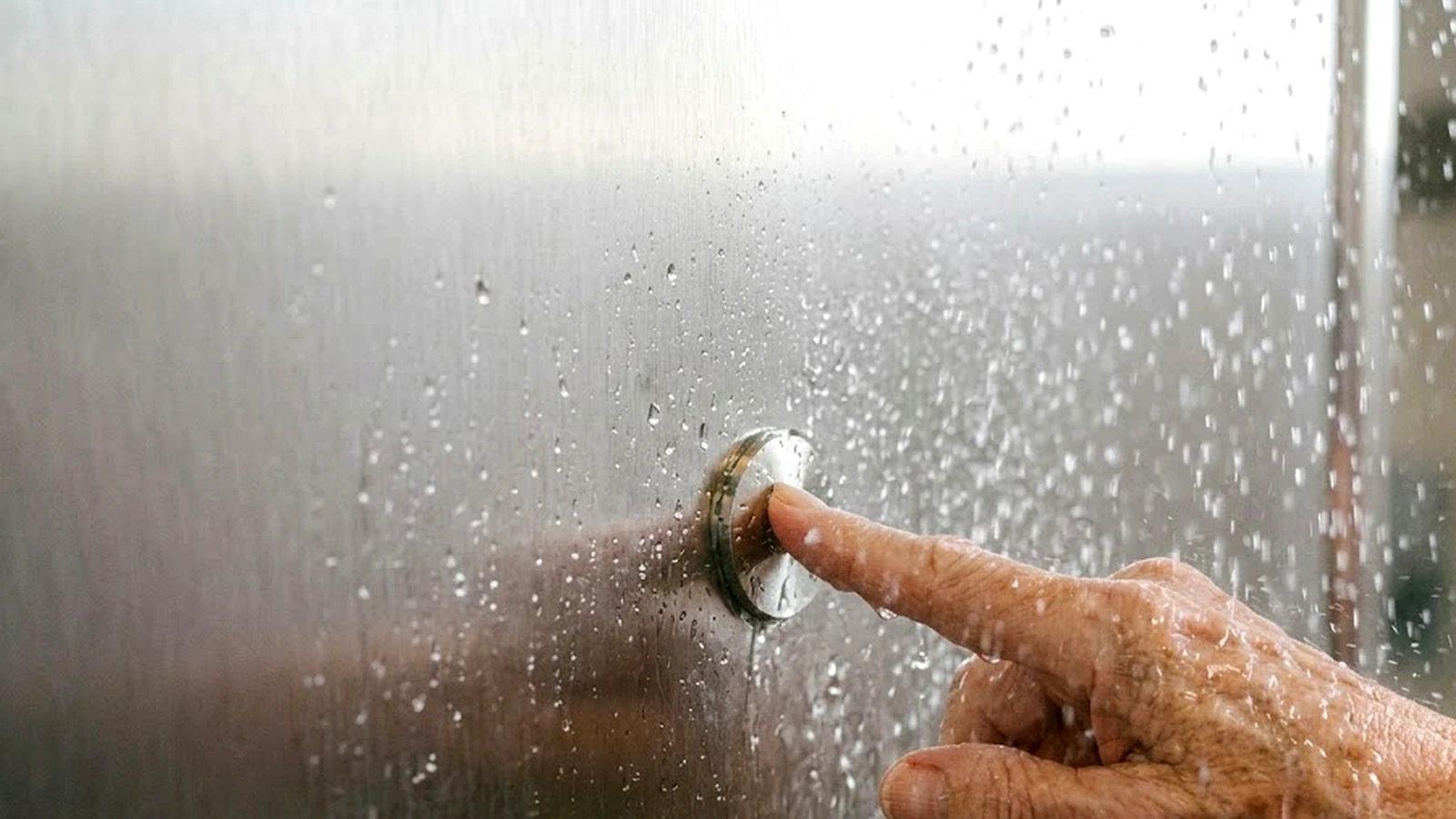 An older person's hand pressing a circular stainless steel control set into a wall panel in a running shower, water on the hand and the panel.